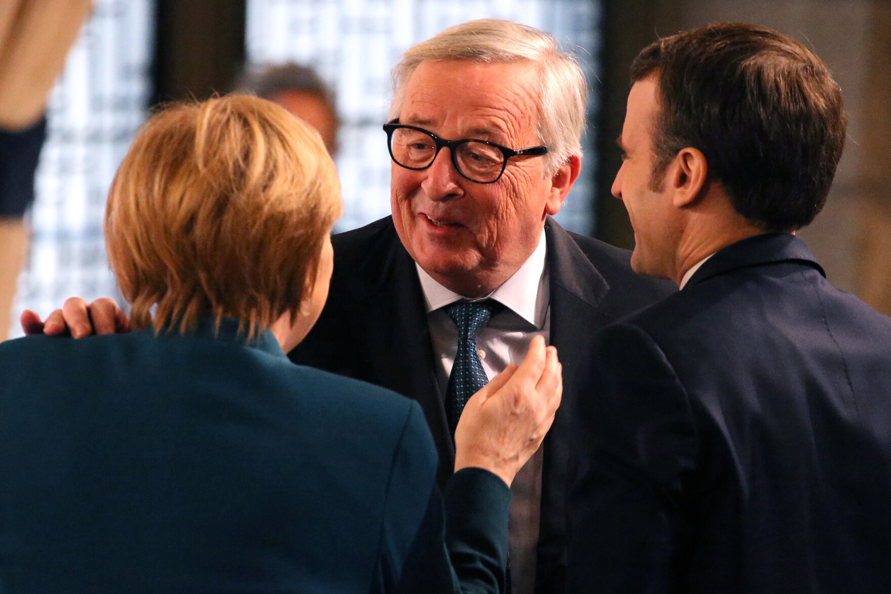 Drei Personen: Angela Merkel und Emmanuel Macron von hinten, Jean-Claude Juncker legt seine Hand auf die Schulter von Frau Merkel und spricht mit ihr.